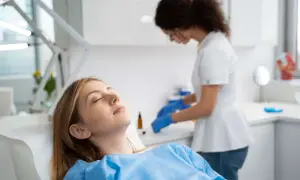 Paziente sdraiata con gli occhi chiusi, forse con un leggero gonfiore guancia dopo anestesia dentista, mentre un'assistente prepara strumenti.