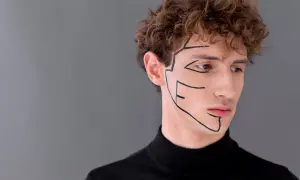 Primo piano di un uomo con riccioli castani, indossa un dolcevita nero. Linee nere disegnate sul viso accentuano l'asimmetria mandibolare.