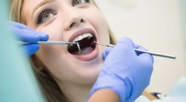 Visita dentistica: una giovane donna con un sorriso smagliante apre la bocca mentre il dentista esamina i suoi denti con strumenti, incluso uno specchietto che riflette il foro mentoniero.