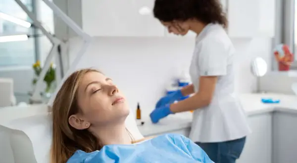 Paziente sdraiata con gli occhi chiusi, forse con un leggero gonfiore guancia dopo anestesia dentista, mentre un'assistente prepara strumenti.