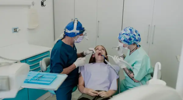 Un dentista con lampada frontale e un assistente eseguono una procedura, forse un'anestesia dentista, su una paziente seduta sulla poltrona.