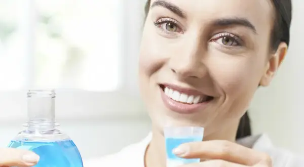 Woman smiling, holding a bottle of blue mouthwash and a cap filled with mouthwash.