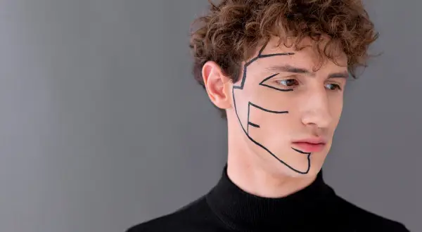 Primo piano di un uomo con riccioli castani, indossa un dolcevita nero. Linee nere disegnate sul viso accentuano l'asimmetria mandibolare.