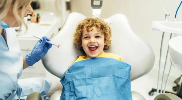 Bambino sorridente dal dentista, pronto per un controllo. Il dentista, con guanti blu, tiene uno specchietto, forse per mostrare cos'è il fluoro e la sua importanza per i denti.