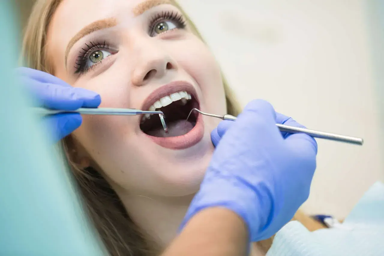 Visita dentistica: una giovane donna con un sorriso smagliante apre la bocca mentre il dentista esamina i suoi denti con strumenti, incluso uno specchietto che riflette il foro mentoniero.
