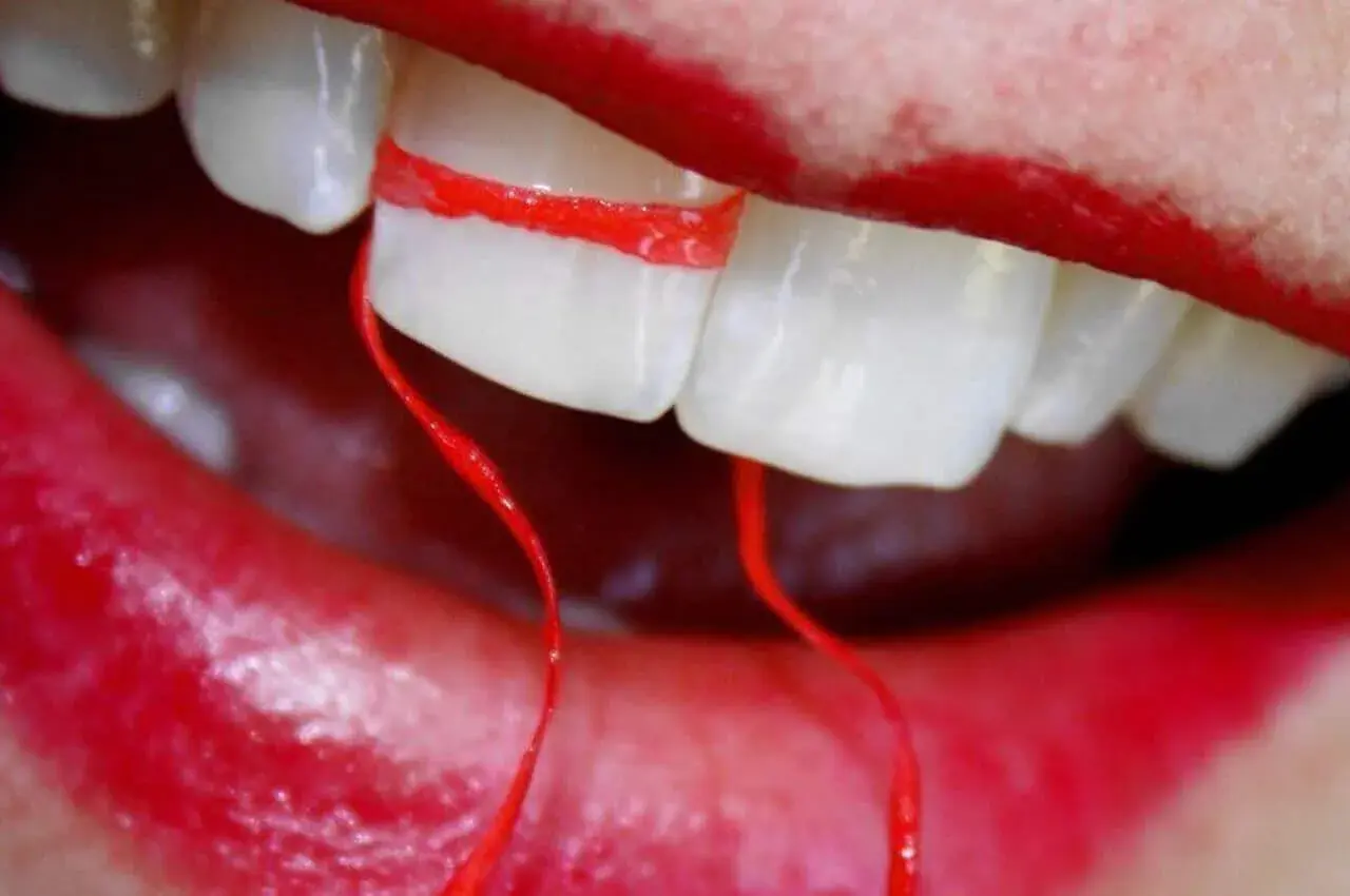 Un filo interdentale rosso avvolge due denti, creando un effetto visivo che sembra allargare i denti. Le labbra rosse incorniciano la scena.