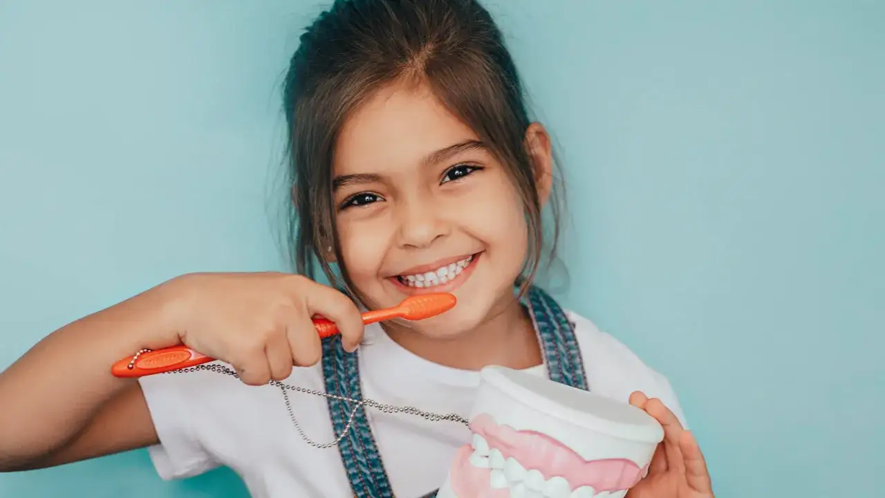Bambina sorridente impara a lavarsi i denti con uno spazzolino arancione e un modello di mascella, promuovendo la fluoro profilassi per denti sani.