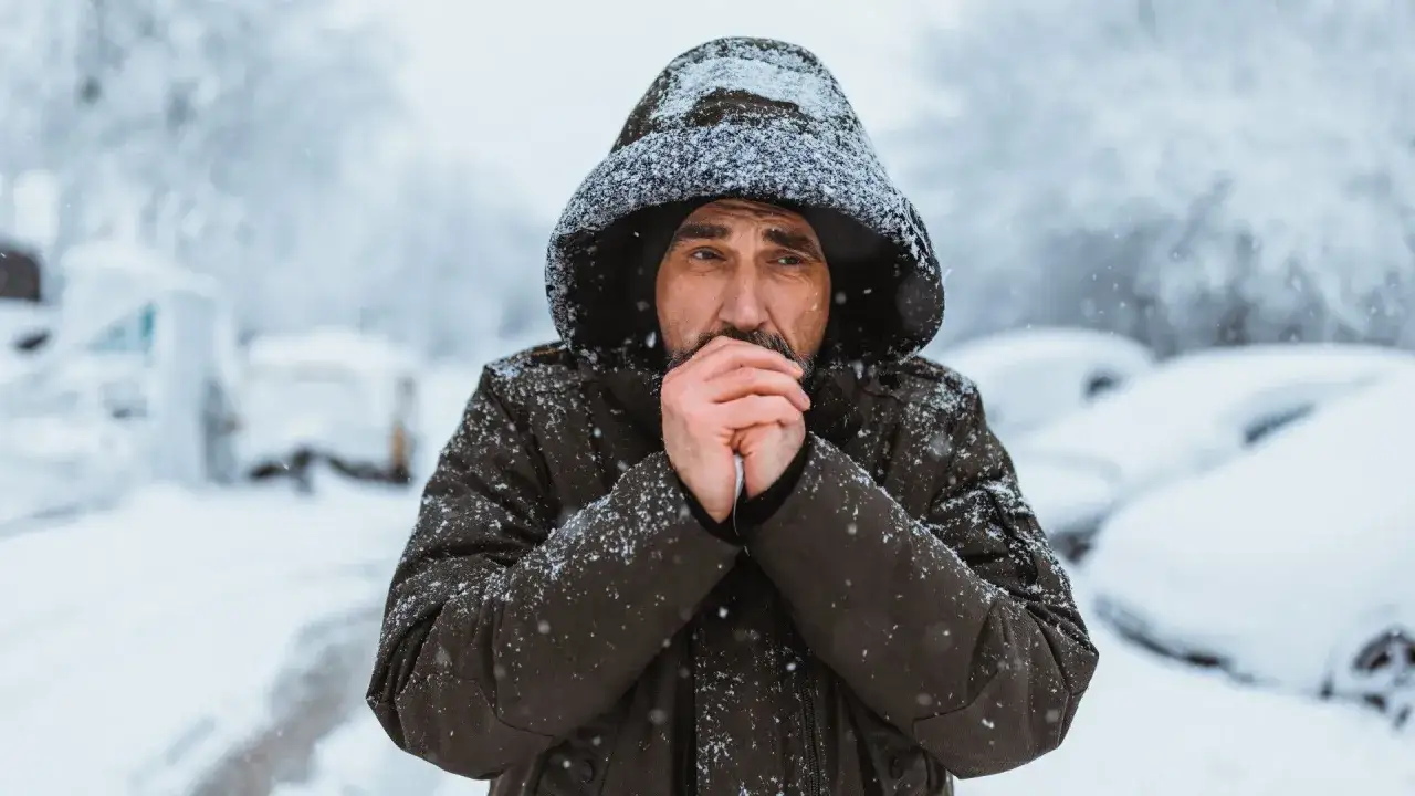 Uomo con cappuccio, mani giunte al viso per scaldarsi, con i denti che battono per il freddo intenso e la neve che cade.
