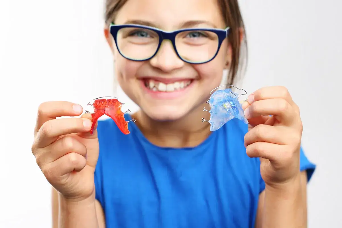 Bambina sorridente con occhiali blu mostra due espansori palatali colorati, uno rosso e uno blu, tenuti tra le dita.