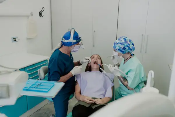 Un dentista con lampada frontale e un assistente eseguono una procedura, forse un'anestesia dentista, su una paziente seduta sulla poltrona.