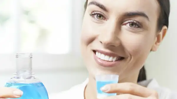 Woman smiling, holding a bottle of blue mouthwash and a cap filled with mouthwash.