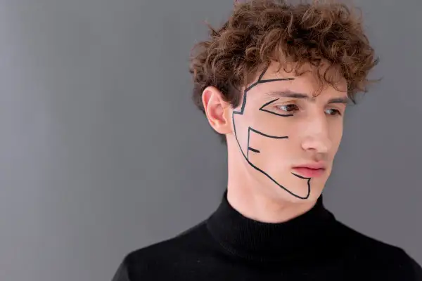 Primo piano di un uomo con riccioli castani, indossa un dolcevita nero. Linee nere disegnate sul viso accentuano l'asimmetria mandibolare.