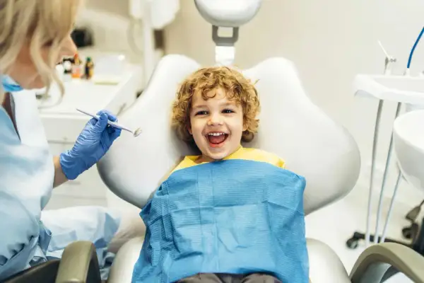 Bambino sorridente dal dentista, pronto per un controllo. Il dentista, con guanti blu, tiene uno specchietto, forse per mostrare cos'è il fluoro e la sua importanza per i denti.
